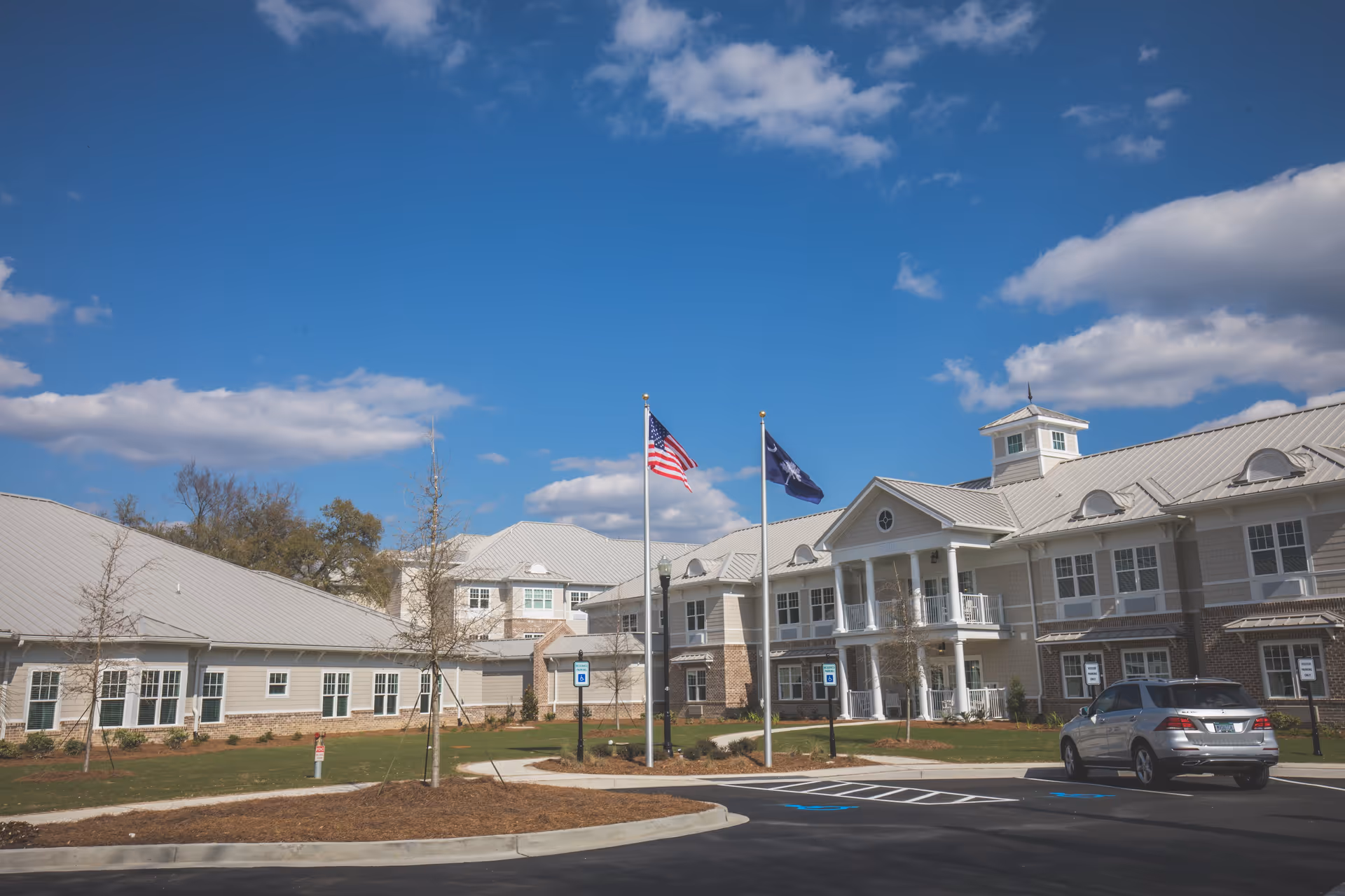 Exterior view of Wellmore of Daniel Island facility showing a large building with white and beige siding, multiple windows, and a metal roof. Two flagpoles with the American flag and a state flag are in front of the building. There is a parking lot with a silver SUV and designated handicapped parking spaces. The sky is clear with some clouds.