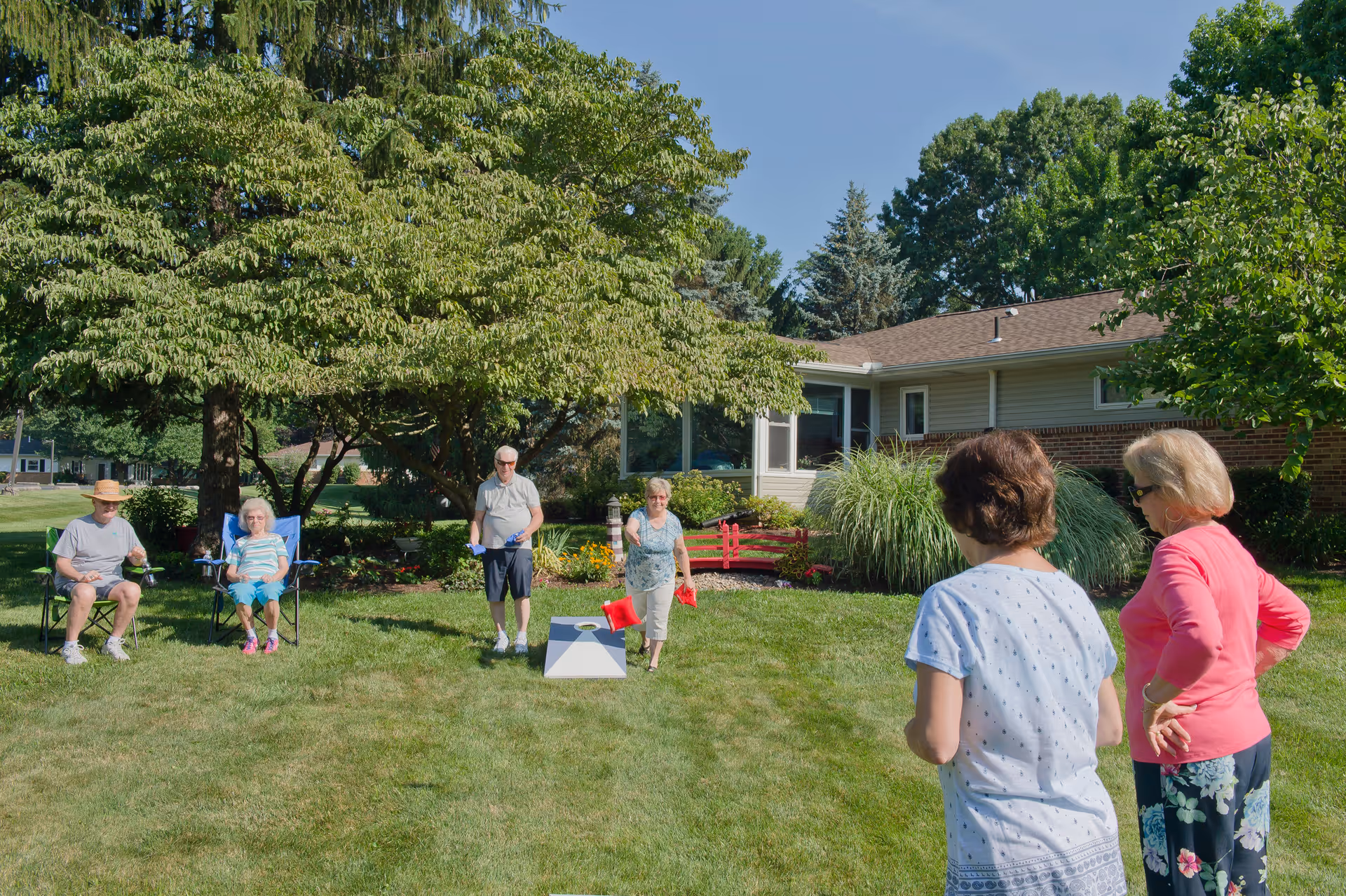 A group of elderly people enjoying an outdoor game of cornhole on a sunny day in a grassy yard with trees and a single-story building in the background.