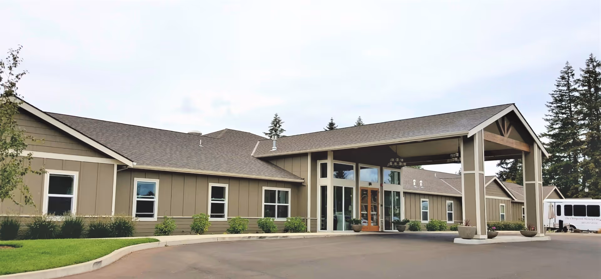 Exterior view of a single-story senior living facility building with a covered entrance and multiple windows. The building is surrounded by a paved driveway, green grass, and some small shrubs. Tall trees are visible in the background under a cloudy sky.