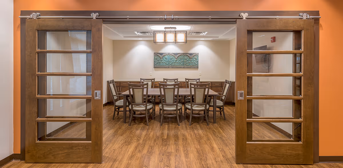 View through wooden sliding glass doors into a dining room with a long table surrounded by chairs and a central pendant light.