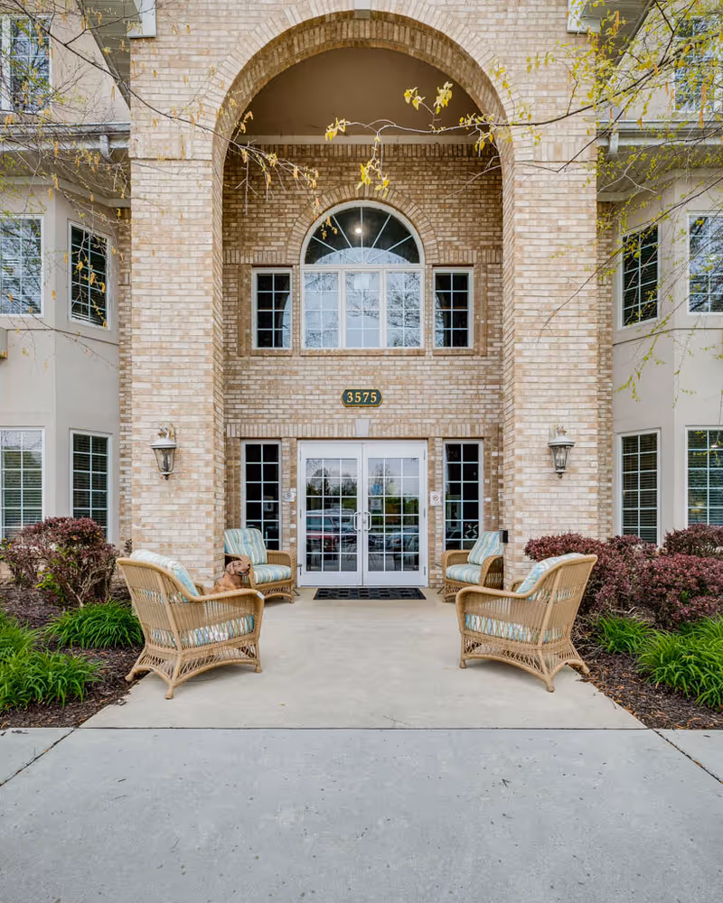 Front entrance of a brick assisted living building with a large arched entry, glass double doors, wicker chairs on the walkway and an address plaque reading "3575".