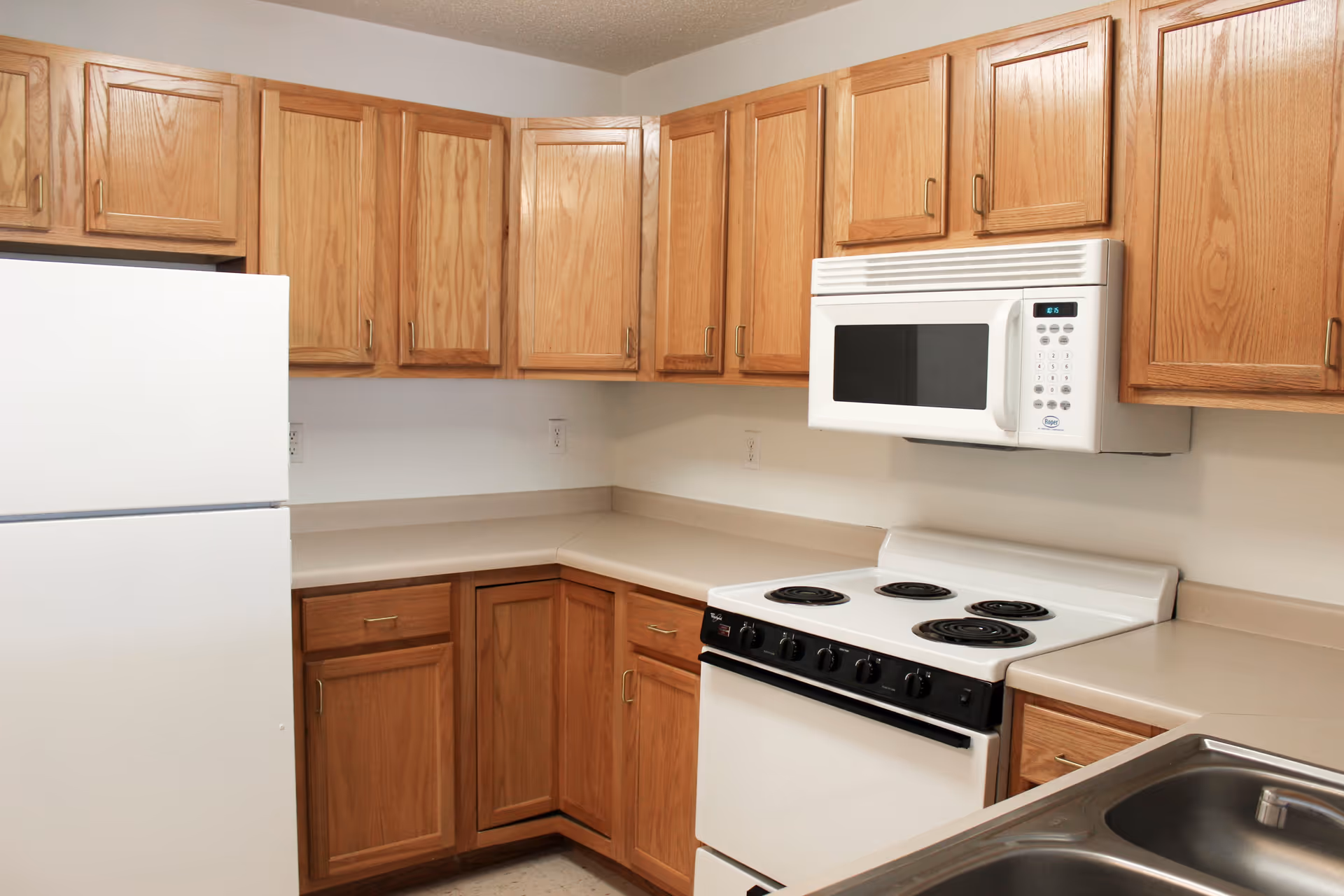A kitchen with wooden cabinets, a white refrigerator, a white electric stove with four coil burners, a white microwave mounted above the stove, beige countertops, and a stainless steel sink in the foreground.