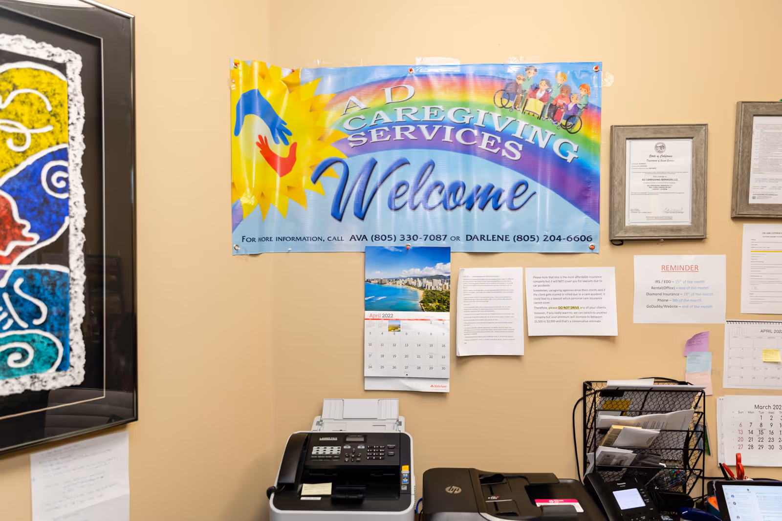 Office wall with A D Caregiving Services welcome banner, calendars, papers, and a printer.