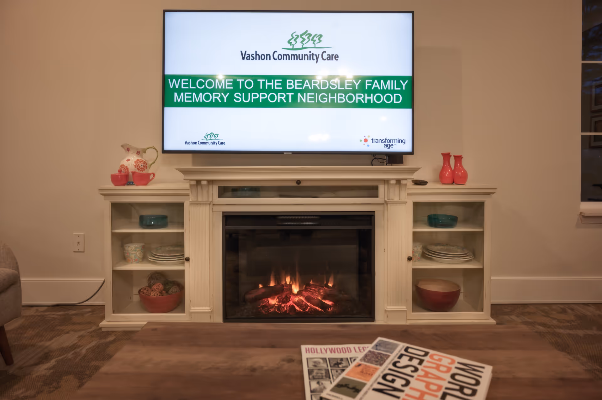 A cozy living room area with a lit fireplace below a mounted flat-screen TV displaying a welcome message for the Beardsley Family Memory Support Neighborhood at Vashon Community Care. The fireplace is flanked by white cabinets with decorative bowls and dishes. A wooden coffee table with magazines is in the foreground.
