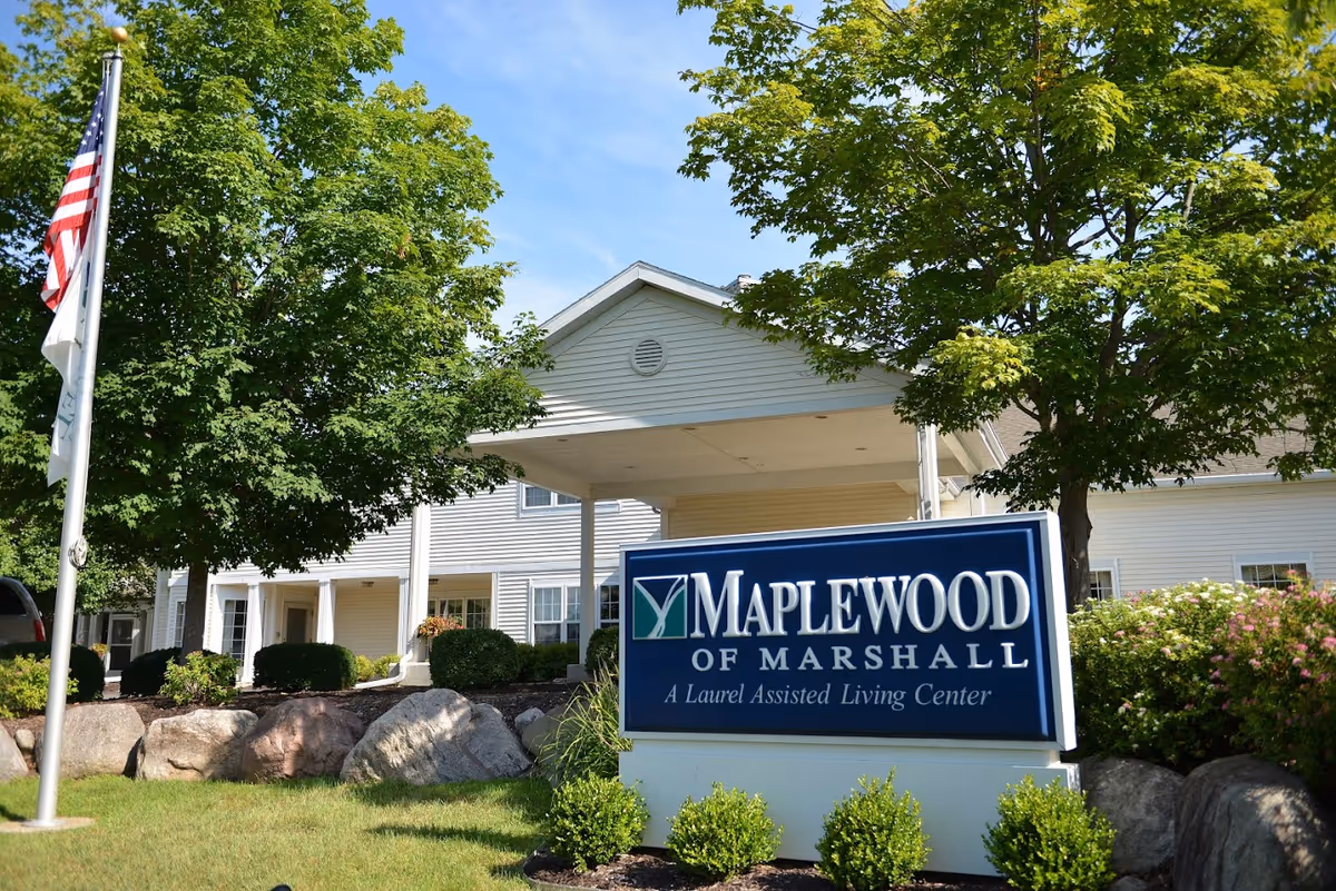 Exterior view of a senior living facility named Maplewood of Marshall with a sign in front that reads 'Maplewood of Marshall, A Laurel Assisted Living Center'. The building is white with a covered entrance, surrounded by green trees, bushes, and rocks. An American flag is visible on a flagpole to the left.