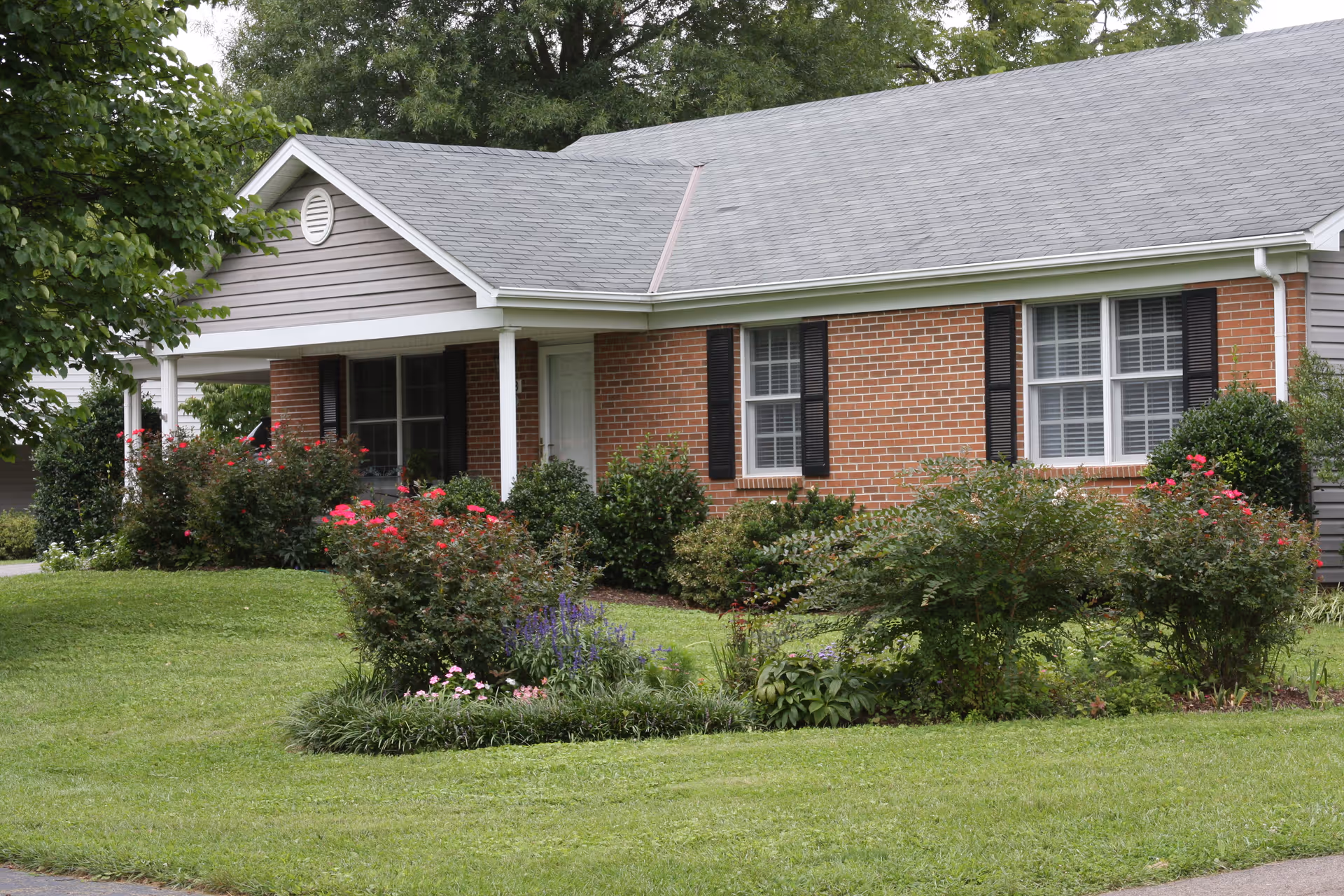 Single-story brick ranch-style house with a covered front porch, manicured lawn, and flowering shrubs.