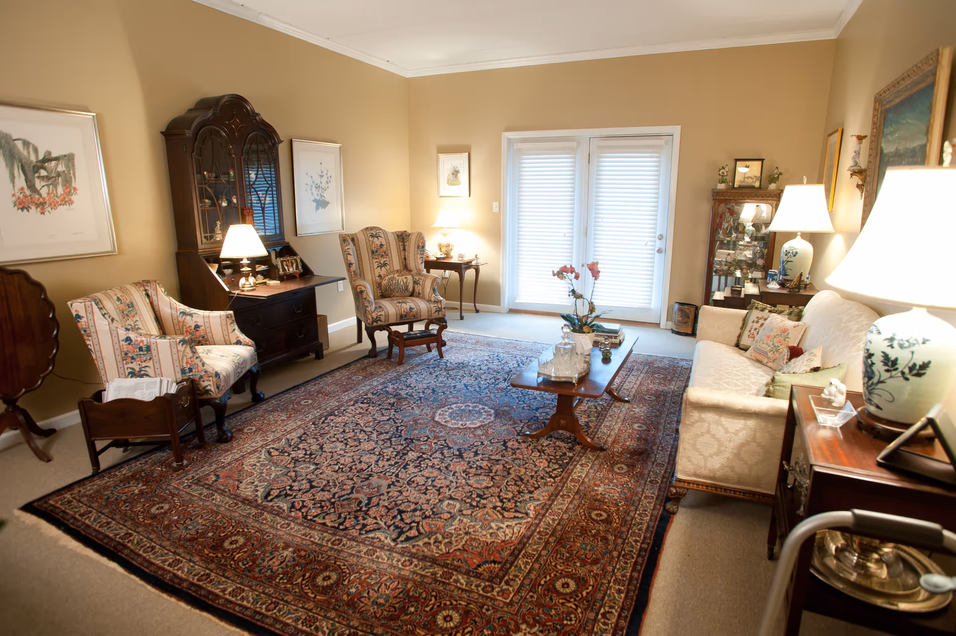 A well-appointed living room with a large patterned area rug, upholstered chairs and sofa, a wooden coffee table, lamps, and French doors with blinds.