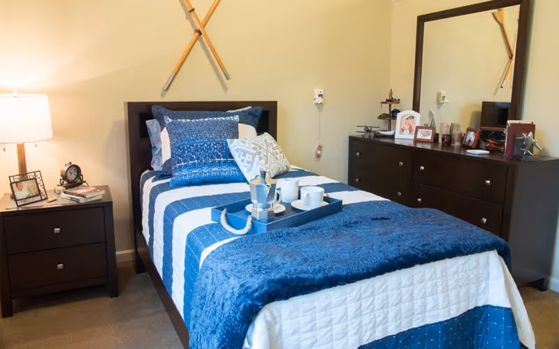 A neatly made single bed with blue-and-white bedding in a furnished bedroom with a nightstand, lamp, dresser, and mirror.