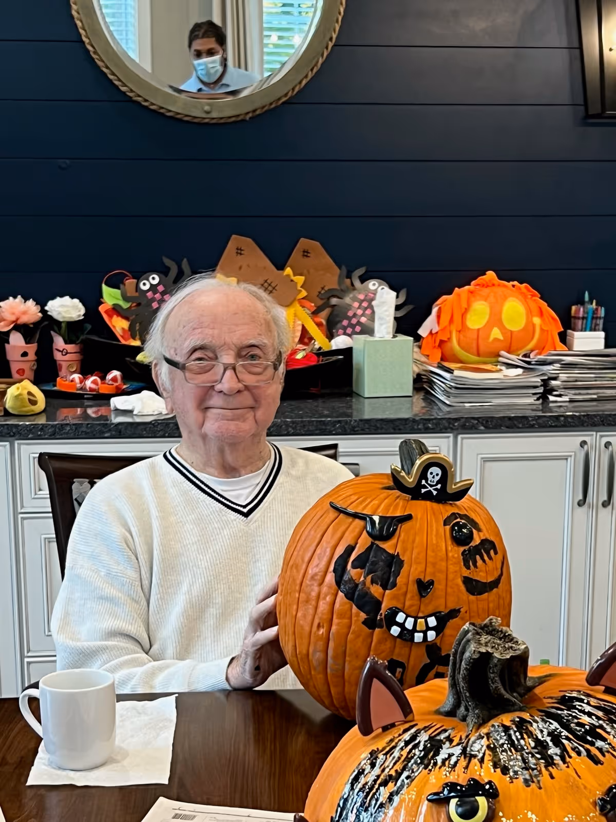 An elderly man wearing glasses and a white sweater sits at a table with decorated Halloween pumpkins in front of him. Behind him is a counter with various Halloween crafts, including paper spiders and a pumpkin decoration. A round mirror on the wall reflects a person wearing a mask.
