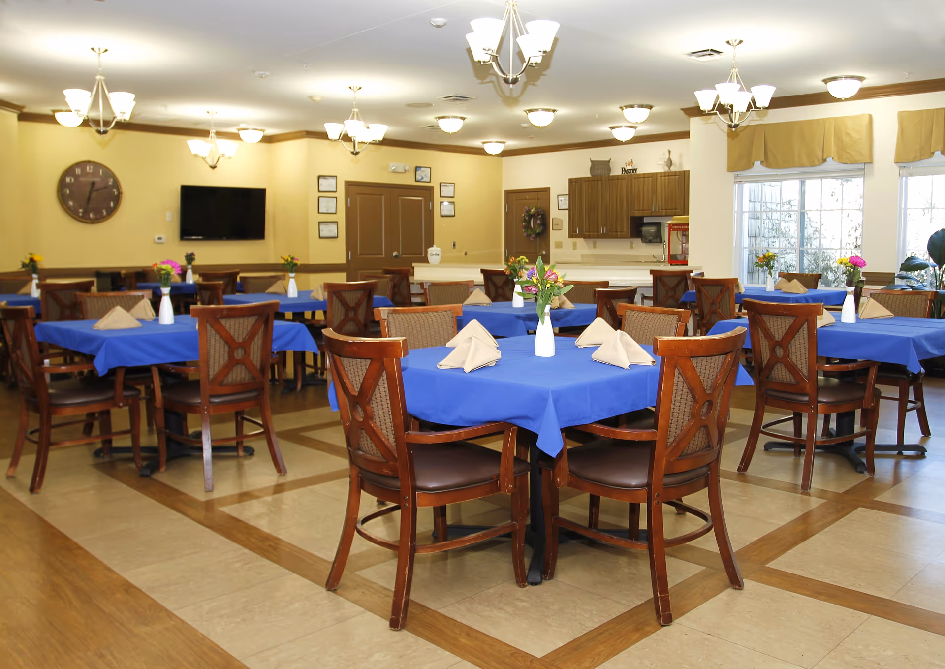 A dining room with multiple wooden tables covered with blue tablecloths, each set with folded beige napkins and white vases holding flowers. The room has wooden chairs around the tables, beige walls, several ceiling light fixtures, a wall clock, a flat-screen TV, and large windows with beige valances letting in natural light.