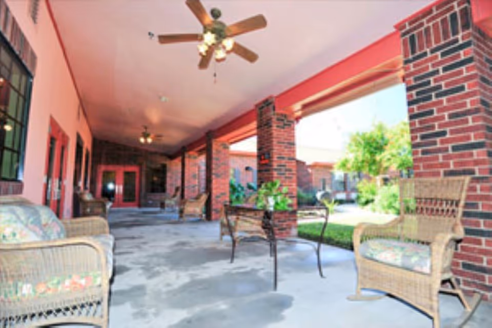 Covered outdoor patio with wicker chairs, brick columns, ceiling fans, and a small table overlooking a courtyard.