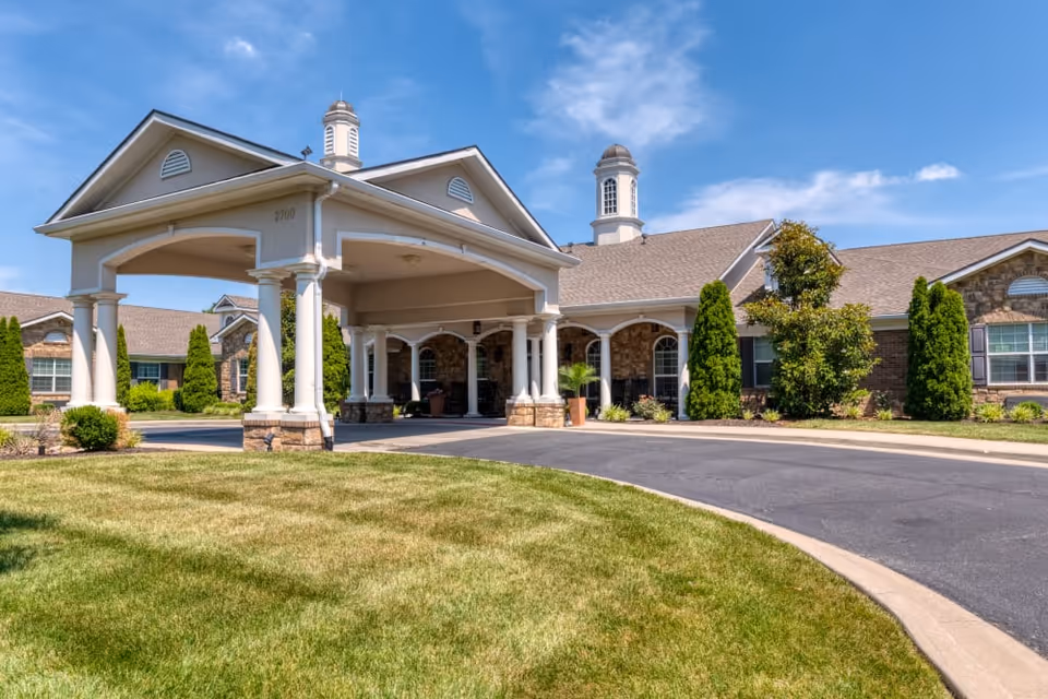 Exterior view of Addington Place of Prairie Village, showing a large covered entrance with white columns, a curved driveway, well-maintained lawn, and a building with stone and brick facade under a blue sky with some clouds.