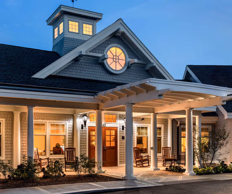 Exterior view of a senior living facility at dusk with warm lights glowing from inside. The building features a covered entrance supported by white columns, rocking chairs on the porch, and a decorative round window above the door. The sky is clear and blue.