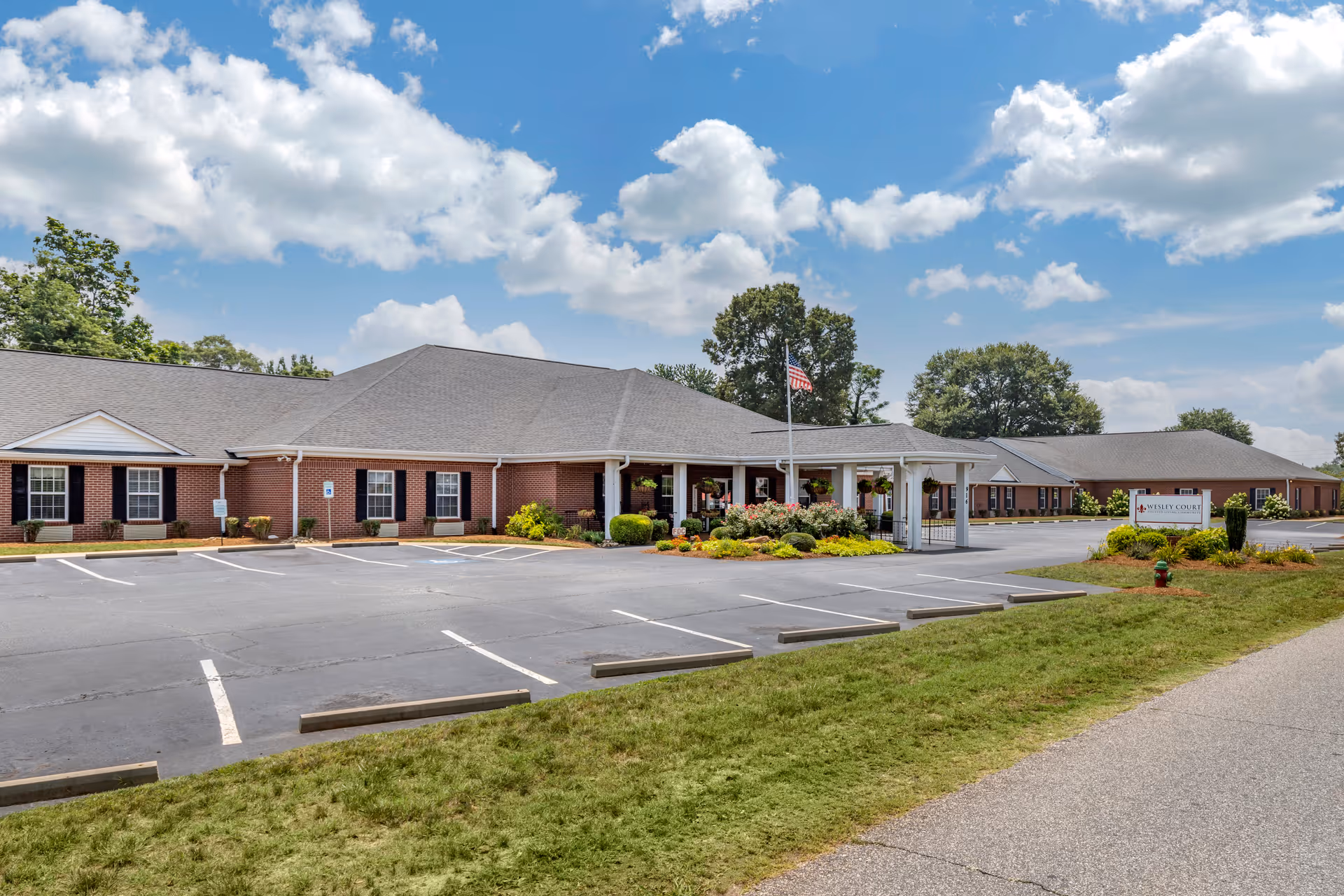 Exterior view of Wesley Court, a single-story brick building with a gray roof, surrounded by a parking lot and landscaped greenery under a partly cloudy blue sky. An American flag is displayed near the entrance, and a sign with the facility's name is visible on the right side near the driveway.