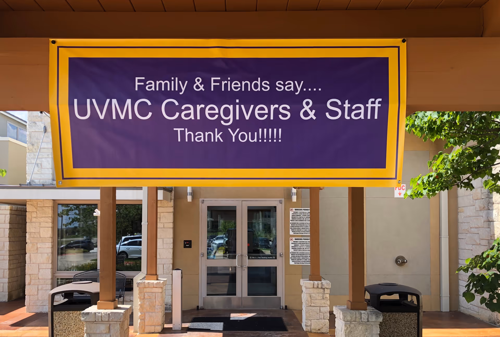 Entrance of a building with a large purple-and-yellow banner reading "Family & Friends say.... UVMC Caregivers & Staff Thank You!!!!!" hung above the double doors.