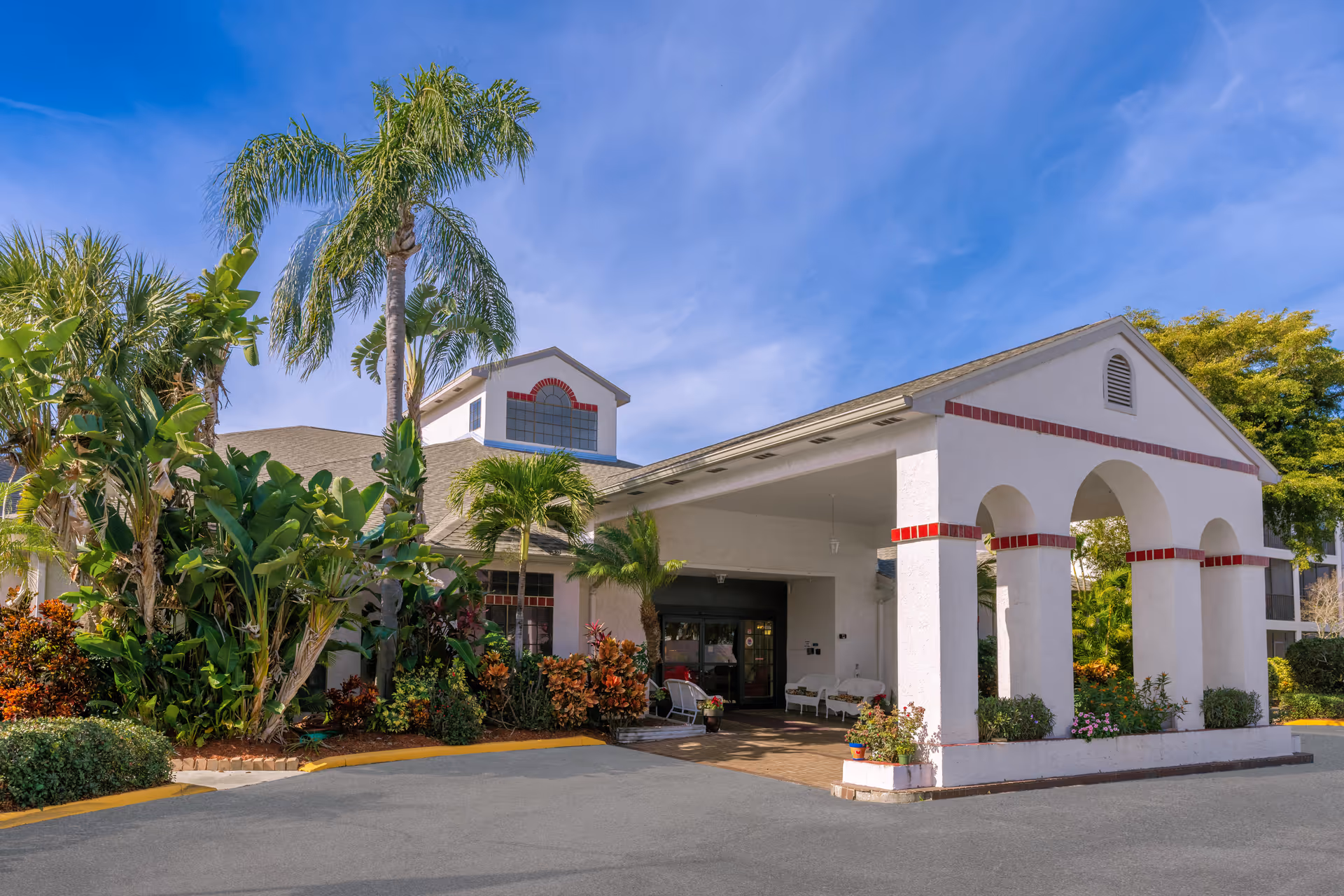 White senior living facility front entrance with arches, driveway, and tropical landscaping.