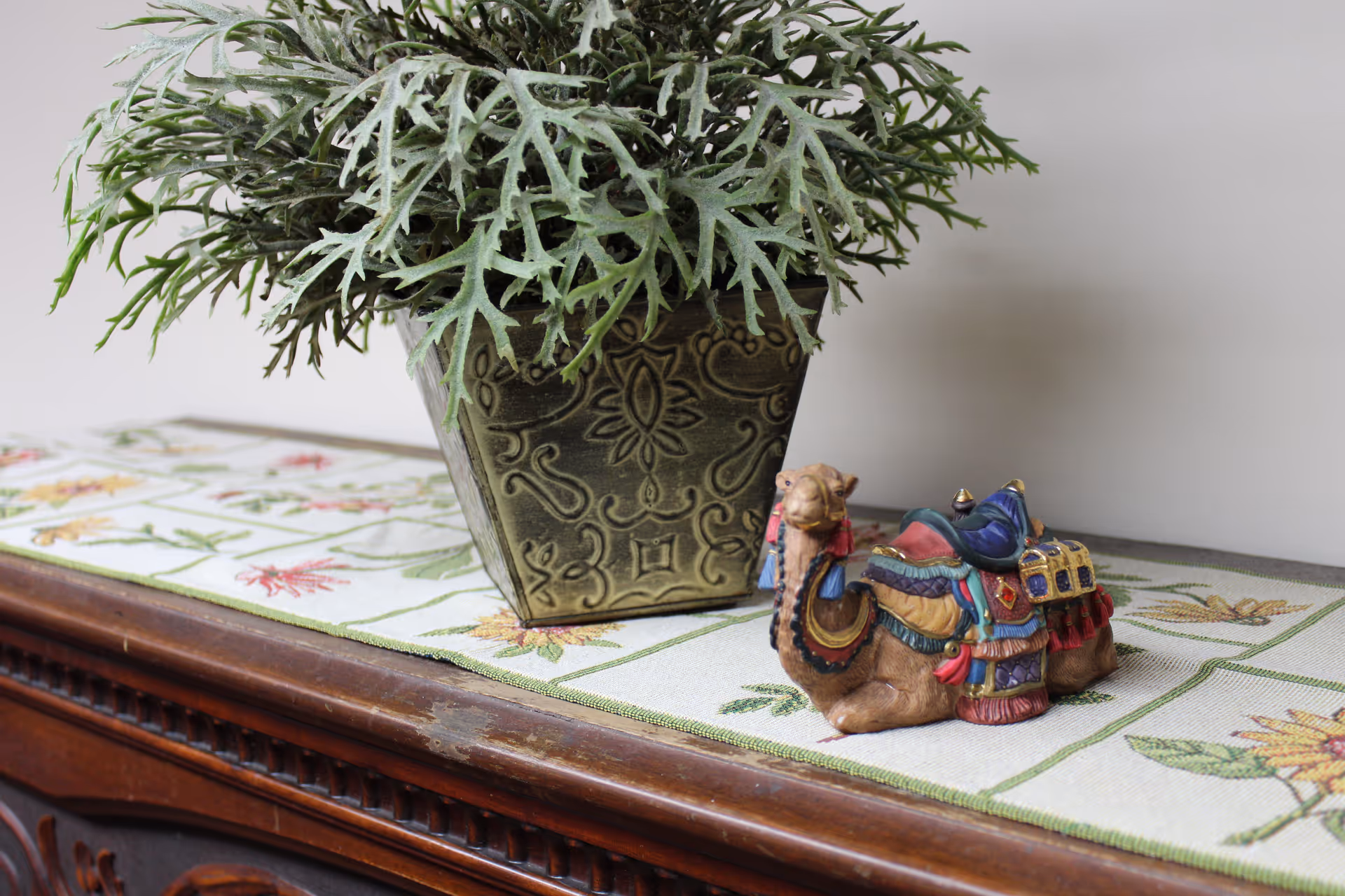 A decorative arrangement on a wooden surface featuring a green potted plant in an ornate square pot and a small colorful camel figurine placed on a floral-patterned table runner.
