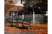 Green metal benches sit on a leaf-strewn path beside a white railing and brick building.