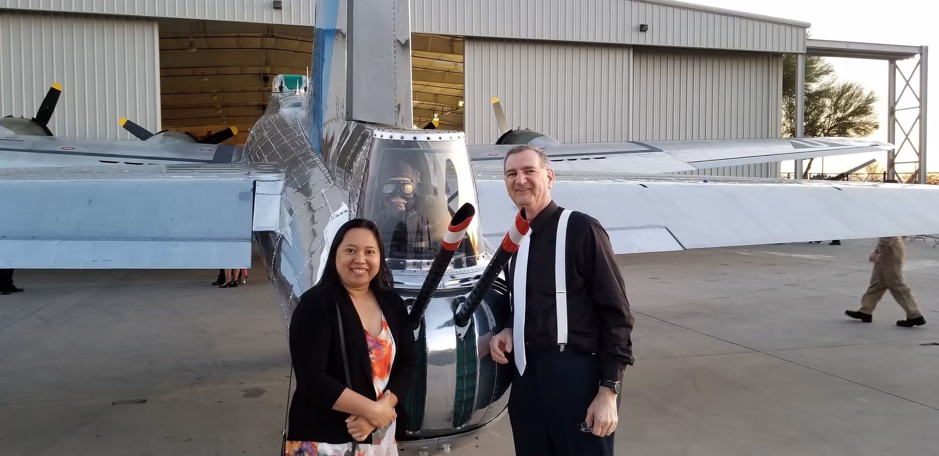 A man and a woman pose beside a silver airplane inside a hangar.