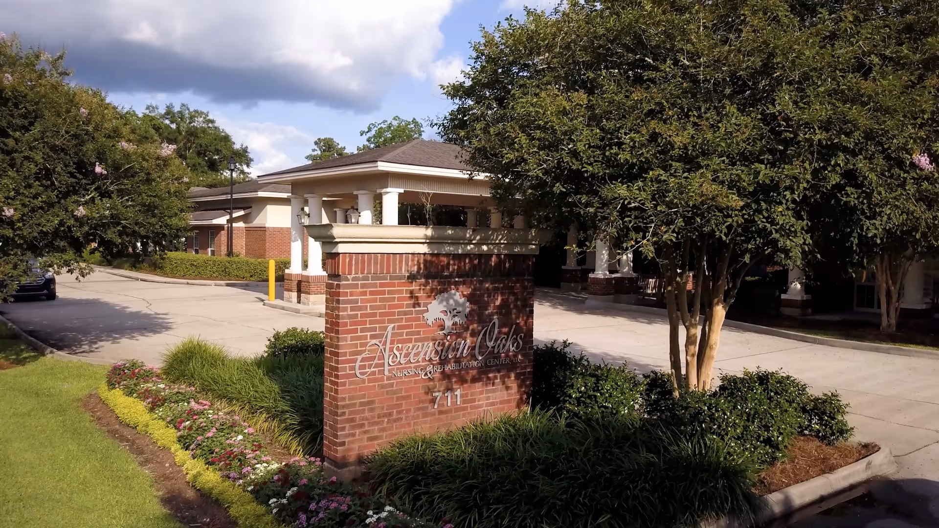 Exterior view of Ascension Oaks Nursing And Rehabilitation Center, LLC showing a brick sign with the facility name and number 711, surrounded by landscaping with flowers and bushes, and a driveway leading to the building entrance under a partly cloudy sky.