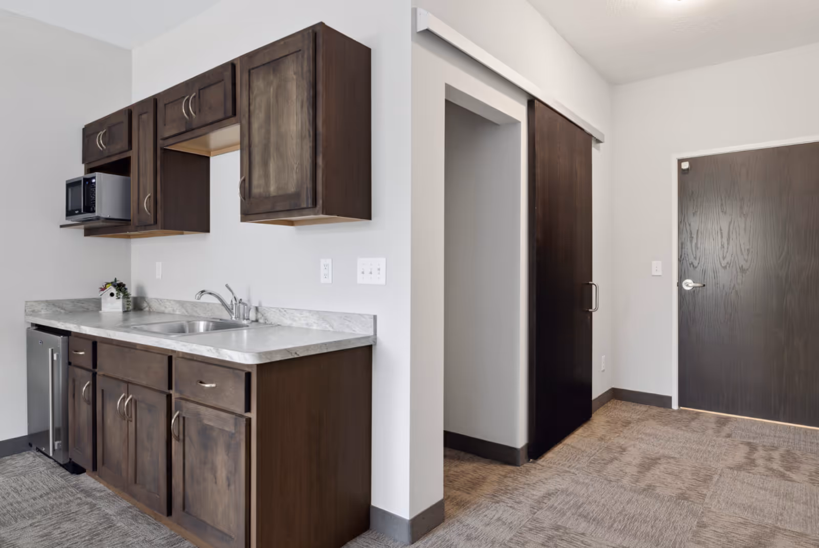 Small kitchenette with dark wood cabinets, marble countertop, sink, microwave, and a sliding door in a residential room.