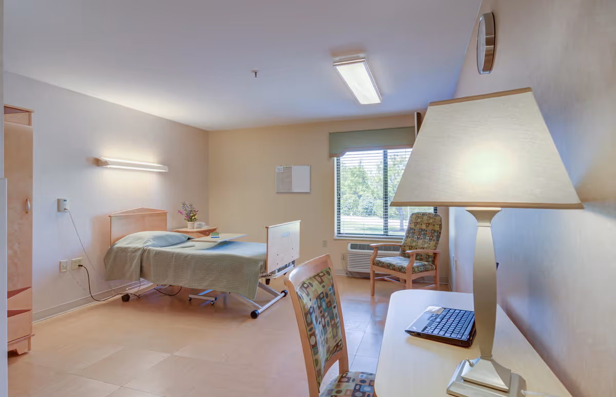 A single-bed senior living bedroom with a desk, chair, and window view.
