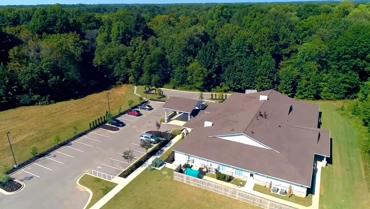 Aerial view of BeeHive Homes of Collierville showing a single-story building with a brown roof surrounded by green trees and grass. There is a parking lot with several cars parked and a covered entrance area.