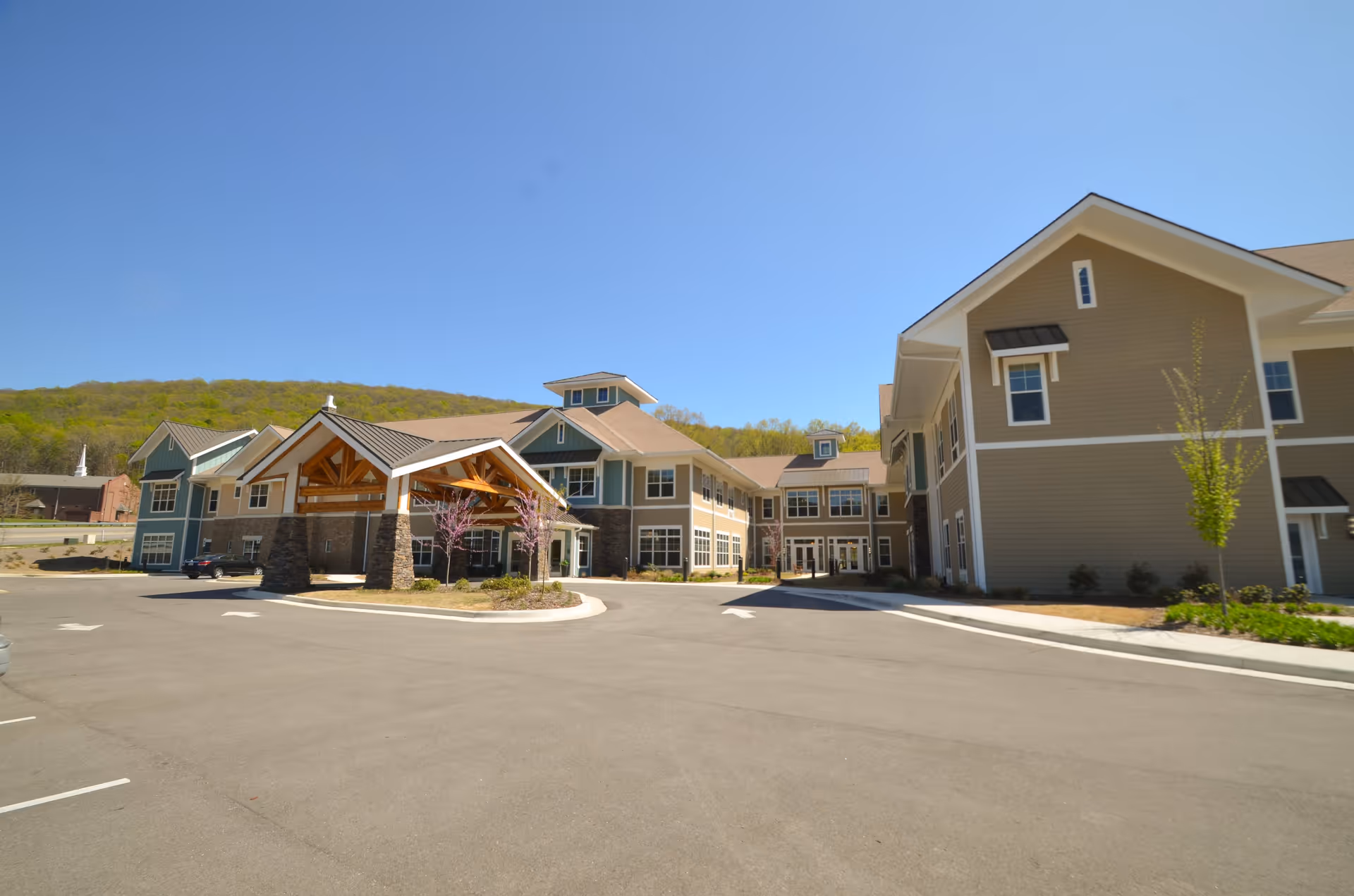 Exterior view of The Goldton at Jones Farm senior living facility on a clear sunny day, showing a large building with multiple windows, a covered entrance supported by stone pillars, a paved driveway, and some landscaping with small trees and shrubs.