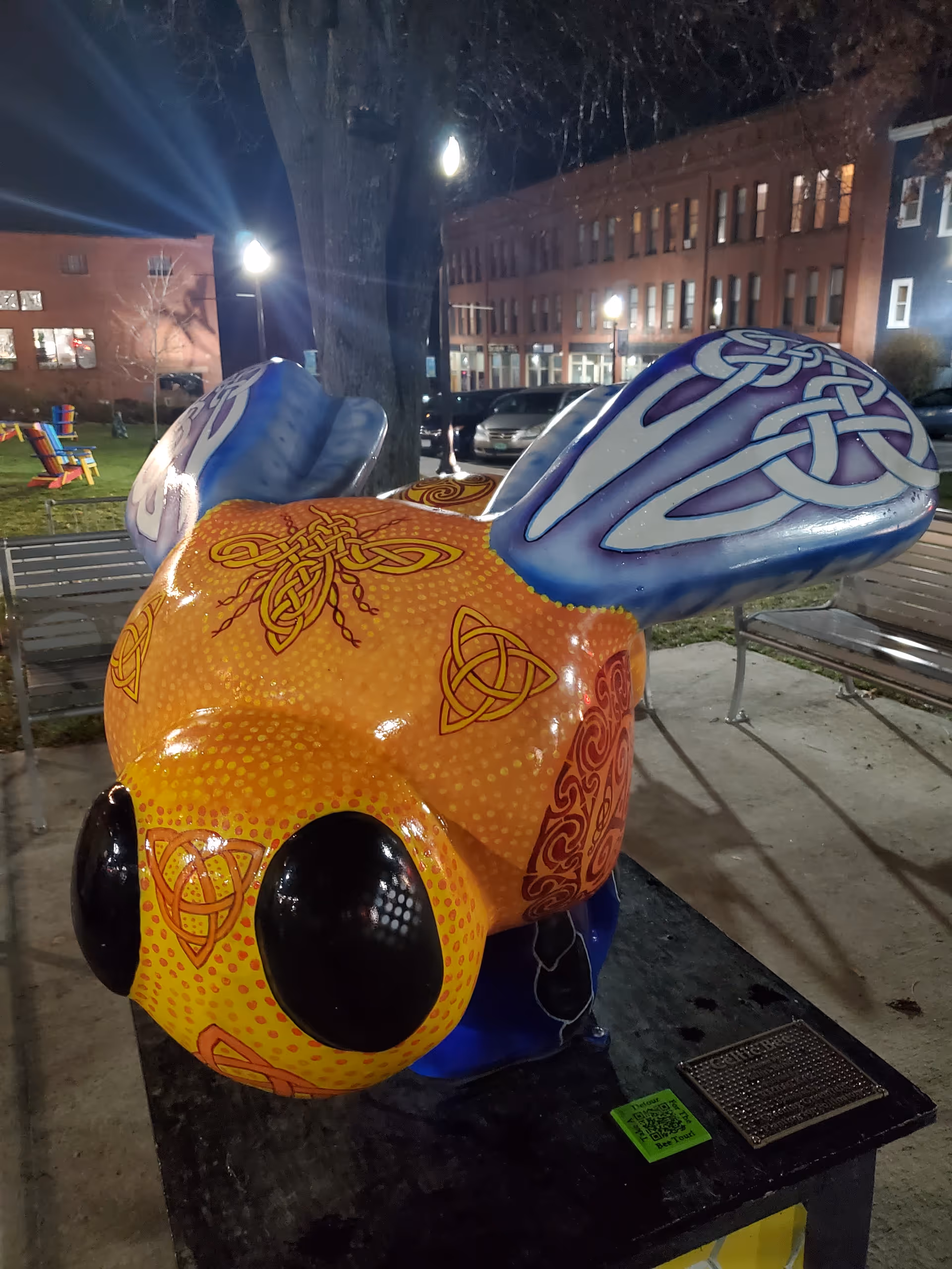 Night view of a colorful bee sculpture in a park near illuminated buildings.
