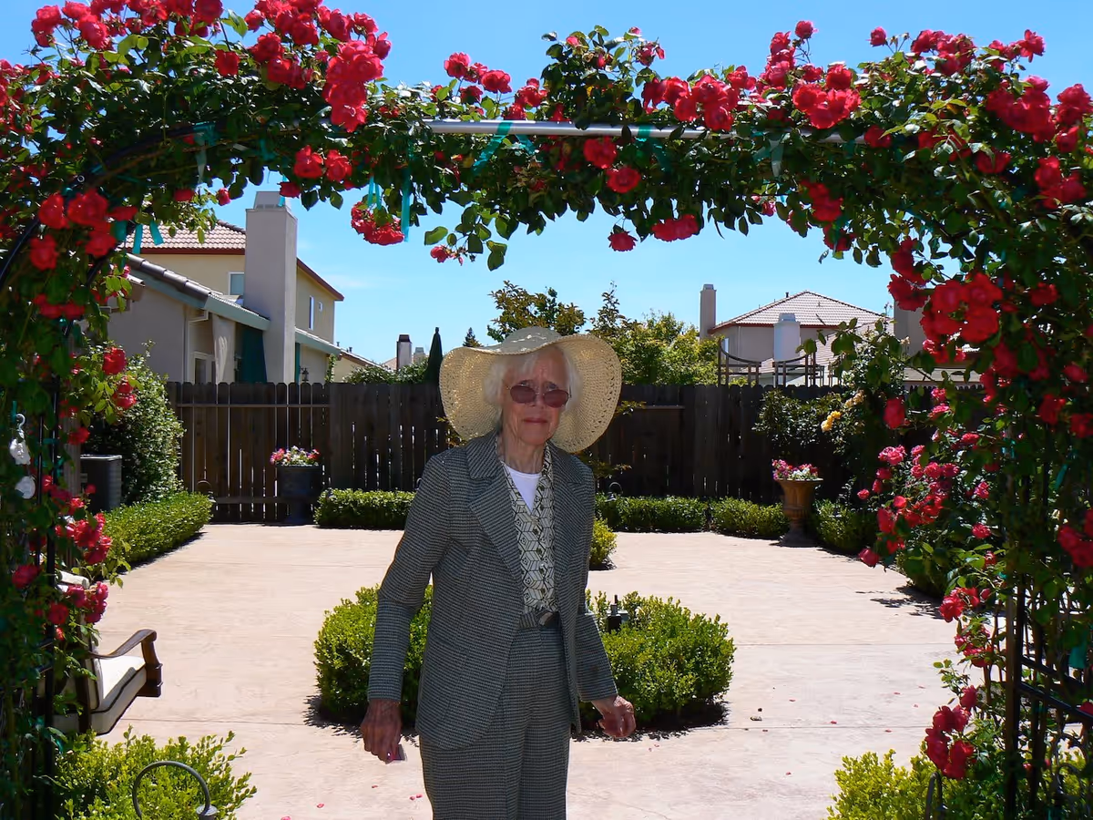 An elderly woman wearing a wide-brimmed hat, sunglasses, and a checkered suit stands under a garden arch covered with vibrant red flowers in a sunny outdoor courtyard with houses and greenery in the background.