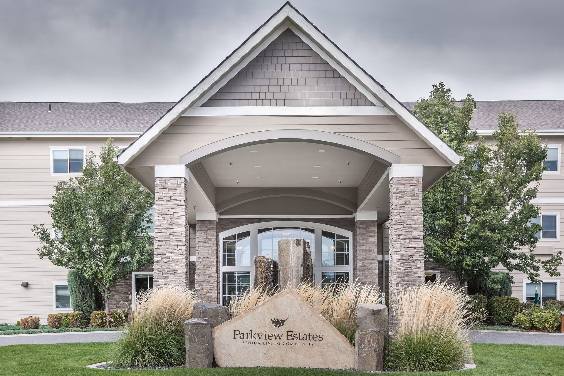 Front entrance of Parkview Estates senior living community featuring a covered driveway with stone pillars, landscaped greenery, and a large stone sign with the facility name.