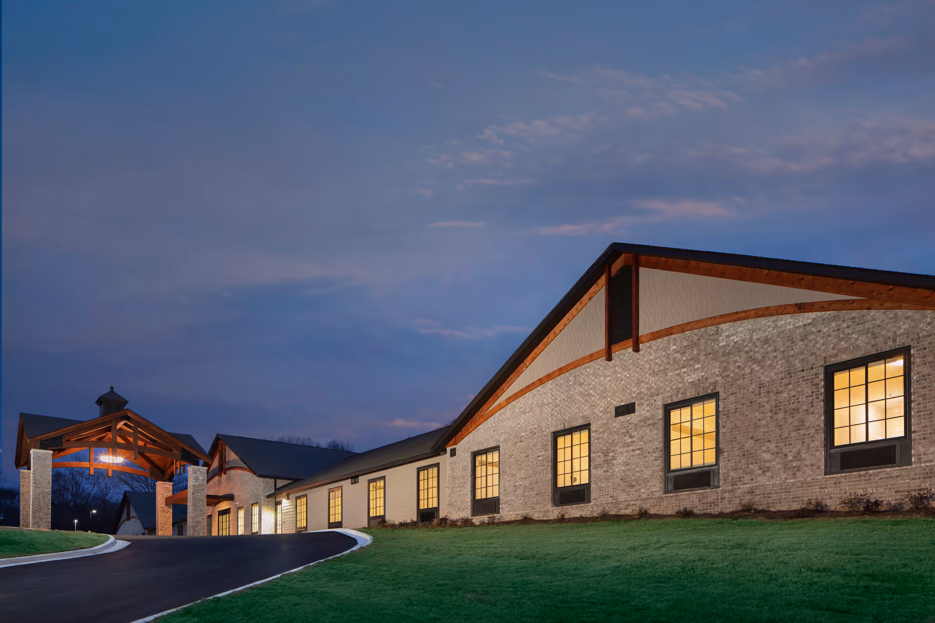 A brick senior living facility exterior at dusk with lit windows and a curved entrance drive.