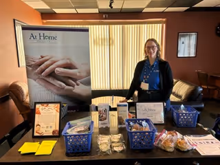 Staff member stands beside a display table of At Home Hospice brochures in a lounge.
