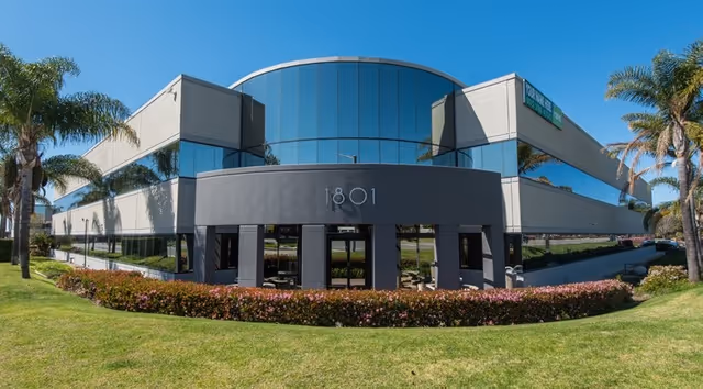 Modern glass-front building with a curved entry and tropical landscaping.