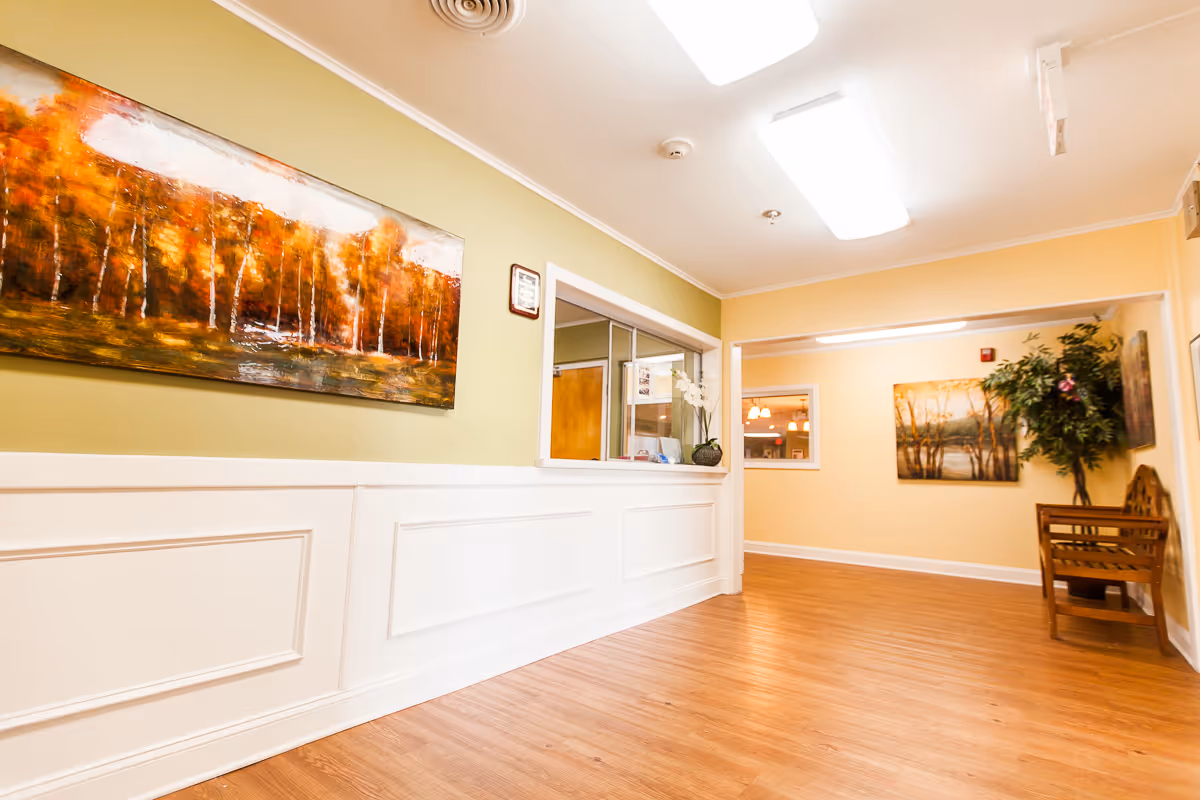 Well-lit interior reception hallway with wood flooring, framed landscape art, a service window, bench seating and a potted plant.