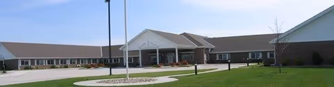 Exterior view of Evergreen Assisted Living facility showing a single-story building with a covered entrance, a flagpole, and a clear sky above.