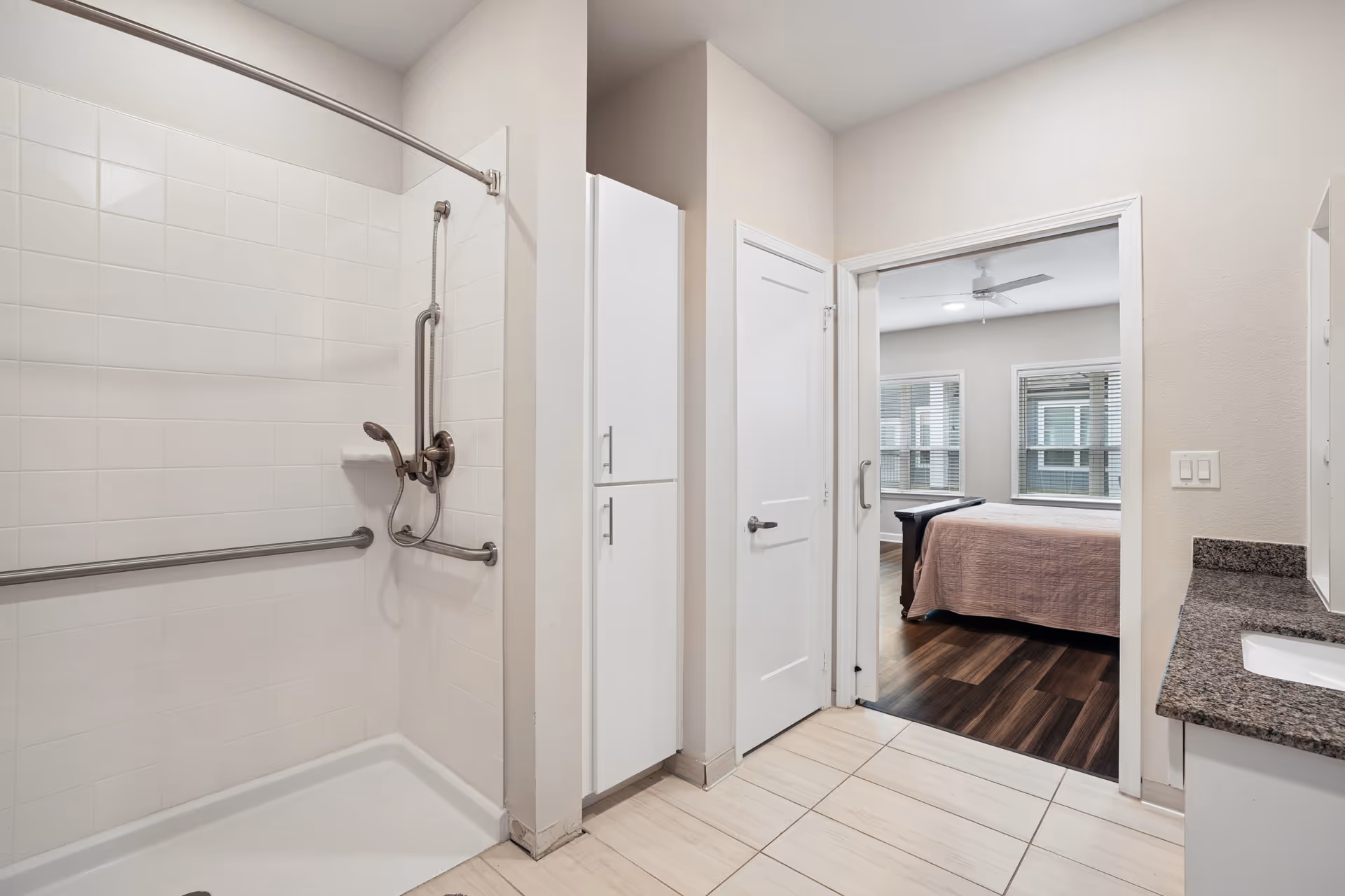 Wheel-in shower with safety bars and doorway to a bedroom in a bright bathroom.