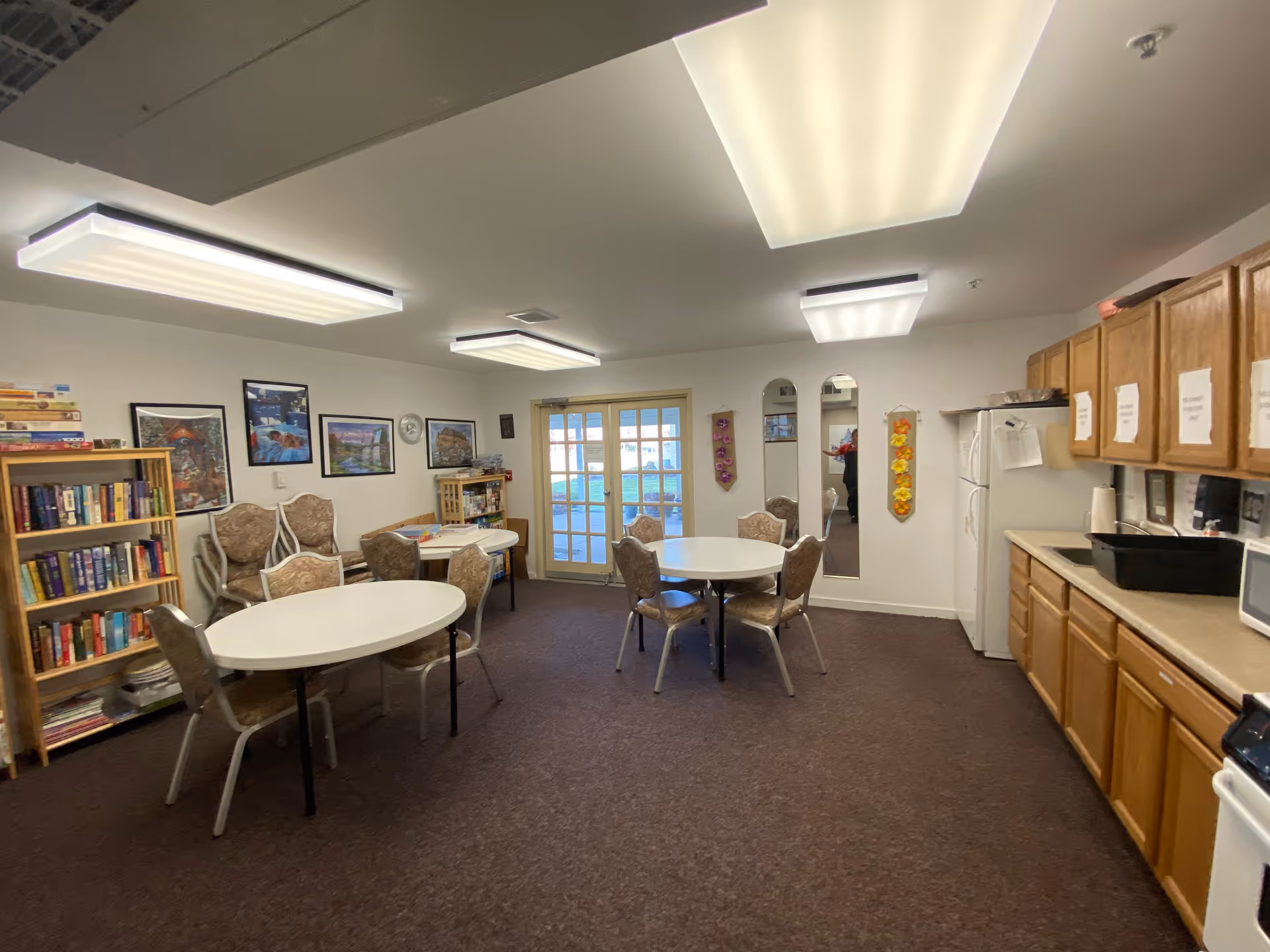 Spacious senior living common area with round tables, chairs, a bookshelf, and kitchenette.