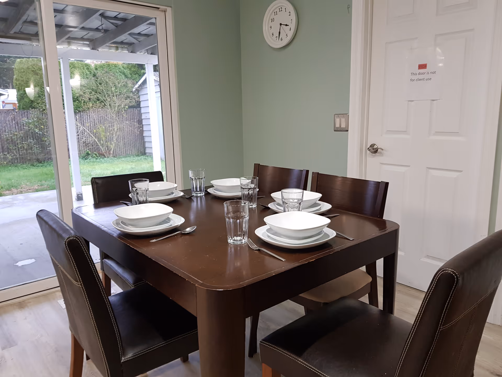 A dining room with a dark wooden table set for six people. Each place setting includes a white bowl, a white plate, a fork, a spoon, and a clear drinking glass. The room has light green walls, a white door with a sign that reads 'This door is not for client use,' and a round wall clock showing the time as 4:25. Sliding glass doors reveal a covered patio and a green backyard with a wooden fence and some bushes.