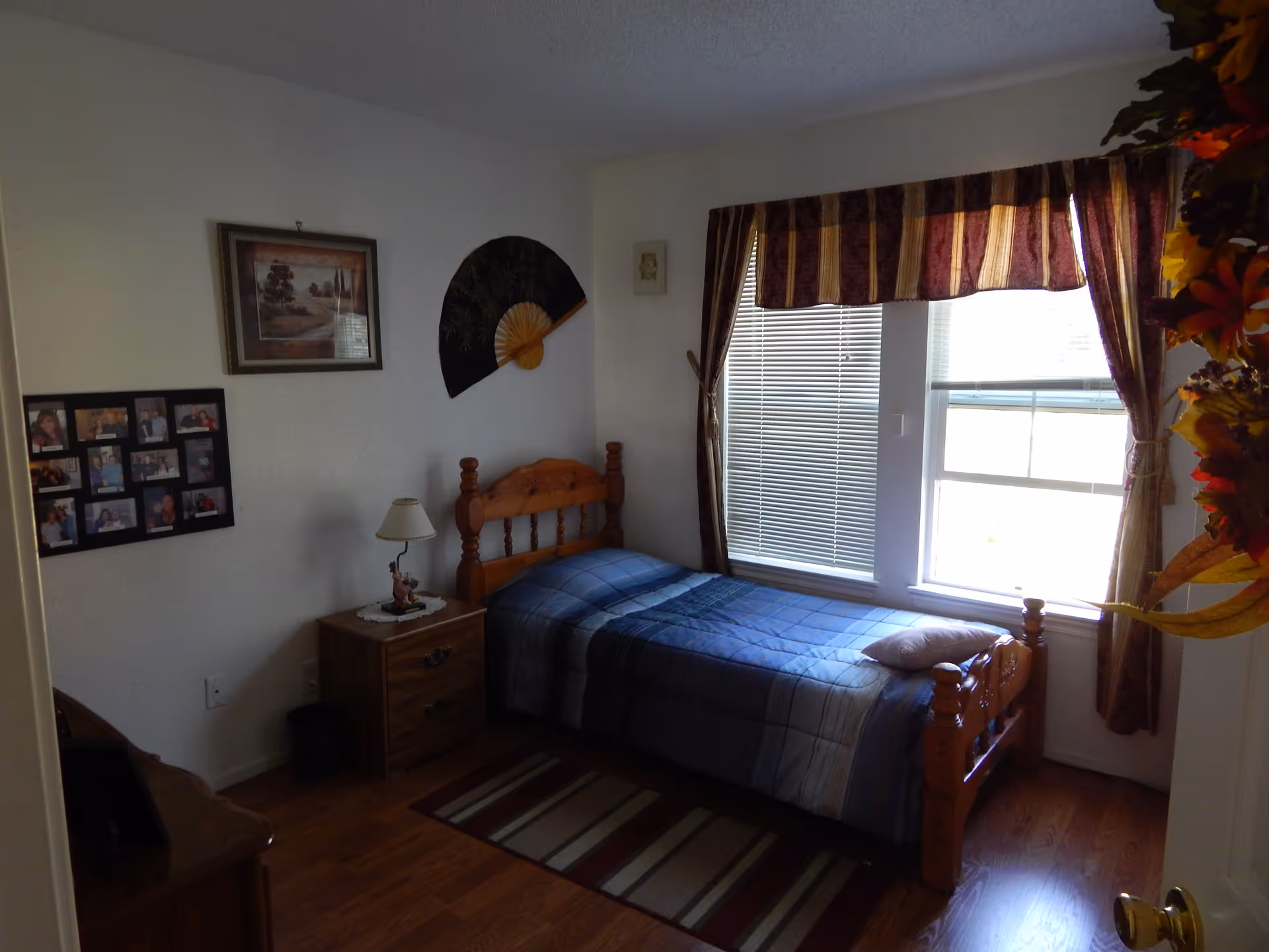 A small bedroom with a single wooden bed covered with a blue plaid blanket and a pillow. Next to the bed is a wooden nightstand with a lamp and a small decorative item. The room has wooden flooring and a striped rug beside the bed. On the wall above the nightstand are framed pictures and a decorative fan. Two windows with blinds and striped curtains allow natural light into the room. A floral wreath is partially visible on the right side near the door.