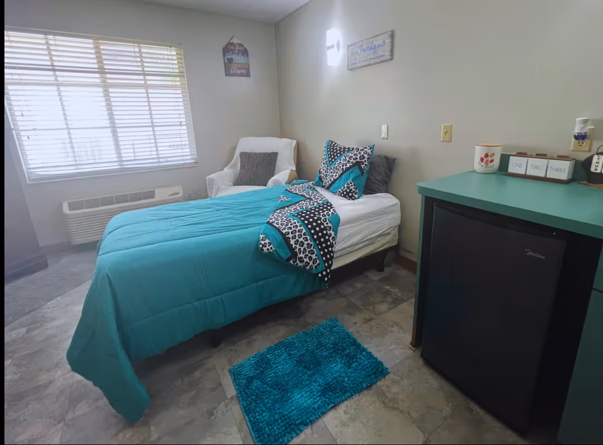 A small, cozy bedroom with a single bed covered in a turquoise and patterned comforter. Next to the bed is a white armchair with a gray pillow. A window with blinds lets in natural light. There is a teal-colored cabinet with a small black refrigerator underneath. The floor is tiled and there is a small turquoise rug beside the bed. The walls are light-colored with two decorative wall hangings.