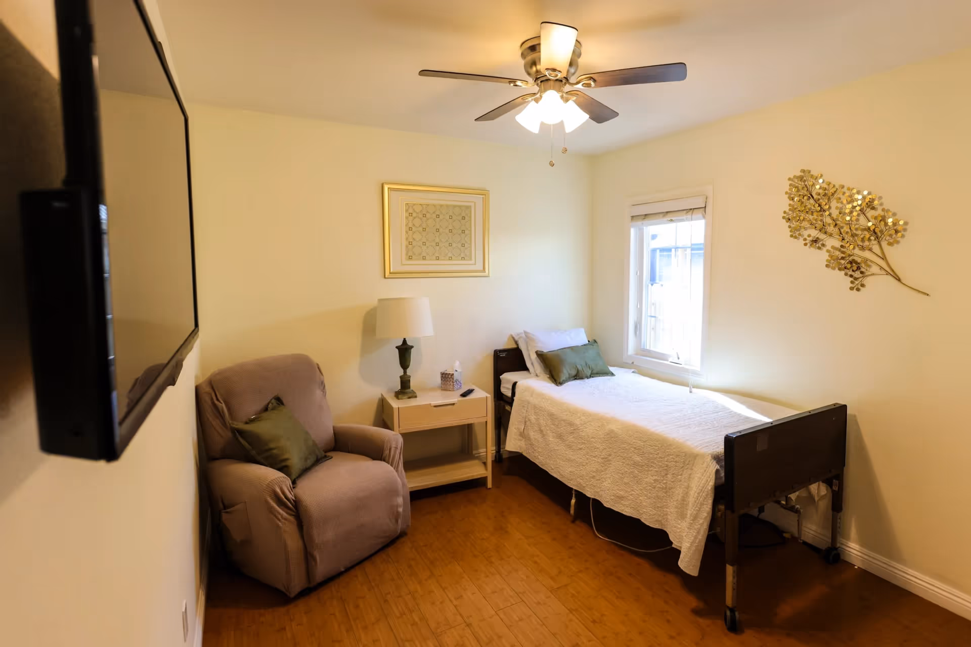 Simple senior bedroom with a single bed, reclining chair, nightstand, ceiling fan and wall-mounted TV.