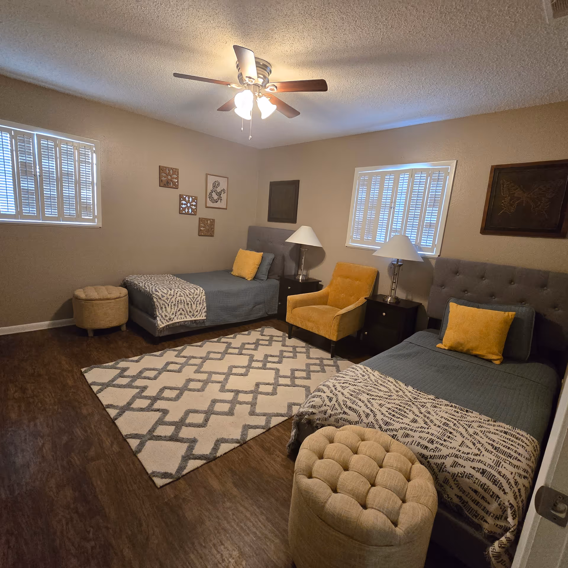 Two twin beds, seating area, nightstands, lamps, ceiling fan, and patterned rug in a cozy bedroom.