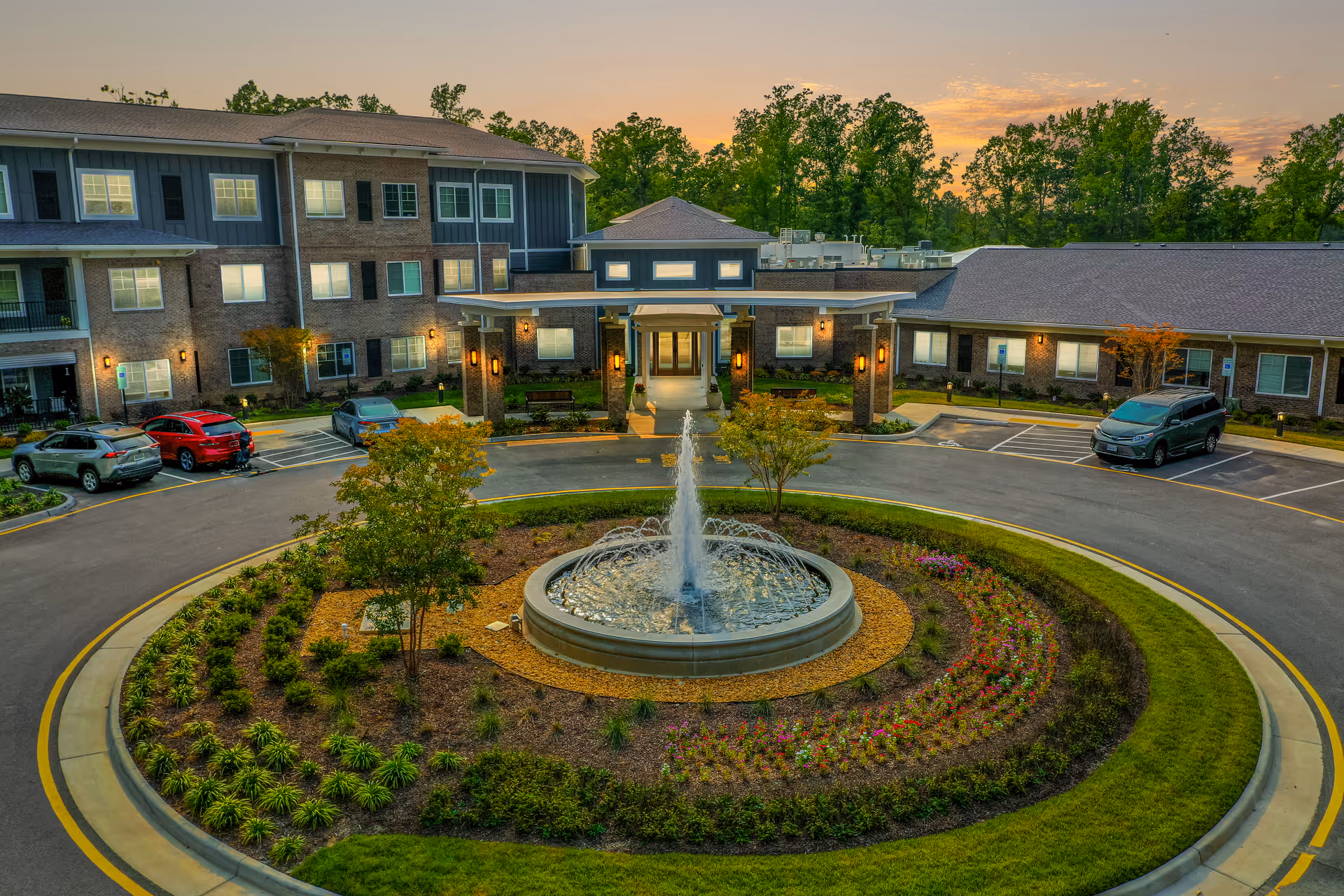 Exterior view of a senior living facility at sunset with a circular driveway featuring a central fountain surrounded by landscaped plants and flowers. The building has multiple windows, a covered entrance with lights, and several parked cars in designated parking spaces.