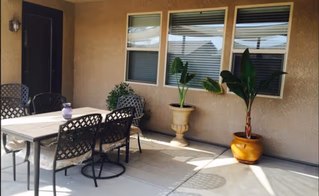 Covered patio area with a dining table and chairs, potted plants, and three windows on the building wall.