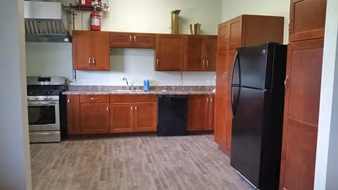 A kitchen with wooden cabinets, a black refrigerator, a black dishwasher, a stainless steel stove with an overhead vent, a countertop with a sink, and a fire extinguisher mounted on the wall. The floor has a wood-like finish.