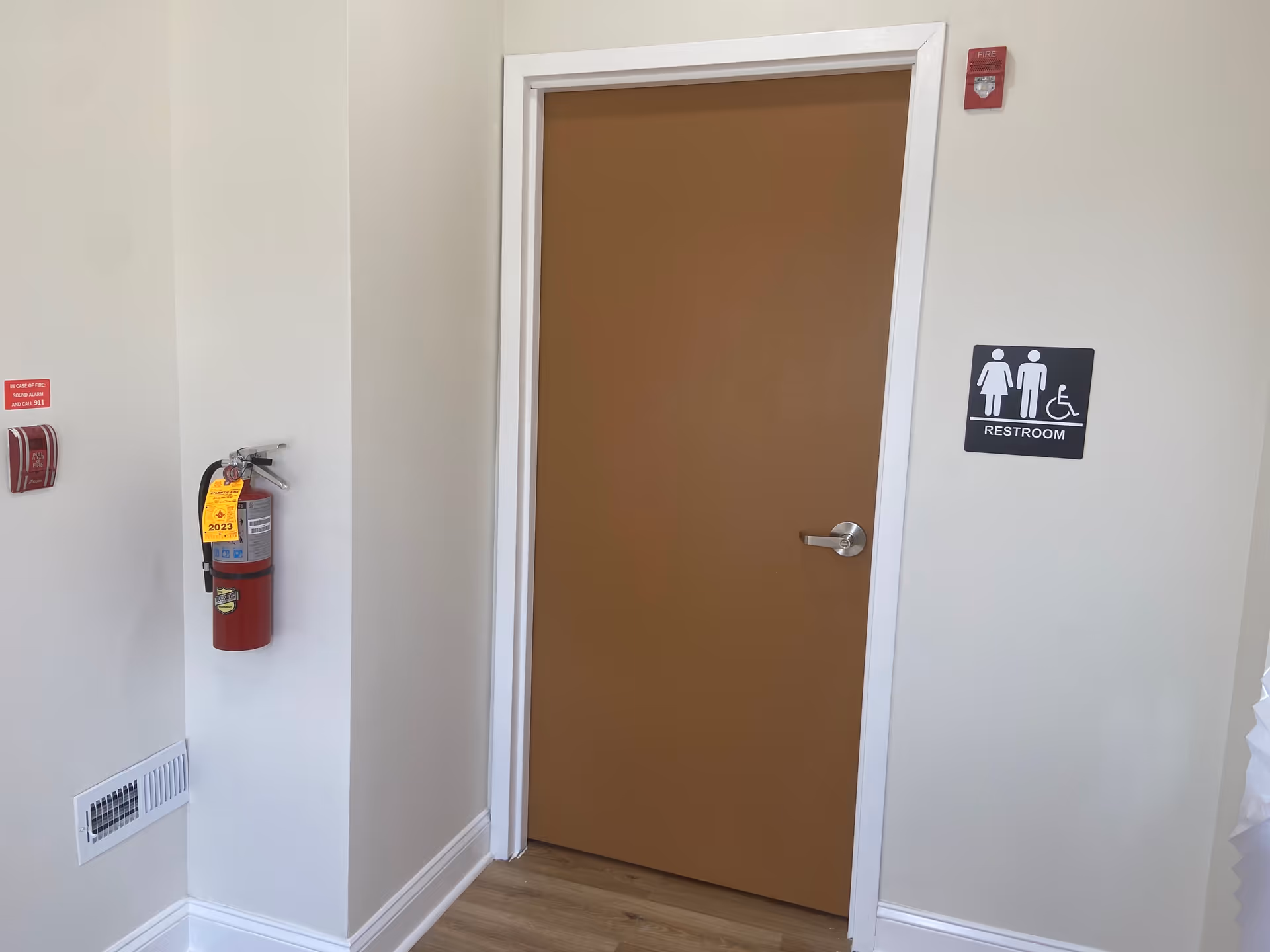 Interior hallway with a brown door, restroom sign, fire extinguisher, and wall vent.