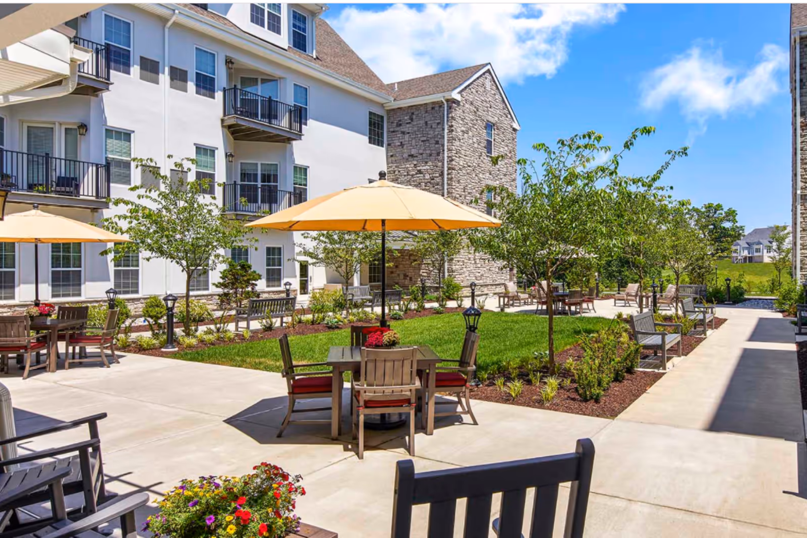 Outdoor courtyard of a senior living facility with tables, chairs, umbrellas, and greenery.
