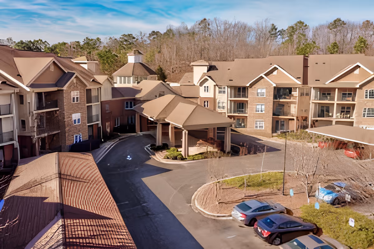 Exterior view of a senior living complex showing the entrance porte-cochere, surrounding multi-story buildings, driveway and parked cars.