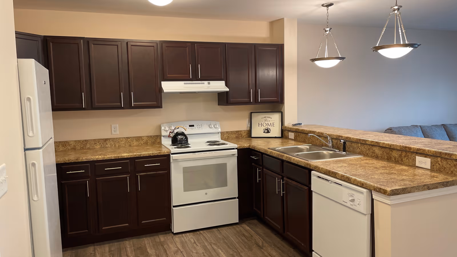 A modern kitchen with dark wood cabinets, a white refrigerator, white electric stove with a kettle on top, a double sink, and a white dishwasher. The countertops are brown with a marbled pattern. Two pendant lights hang above the counter, and a framed sign that says 'Home' is placed on the countertop near the stove. In the background, part of a living room with a gray couch is visible.